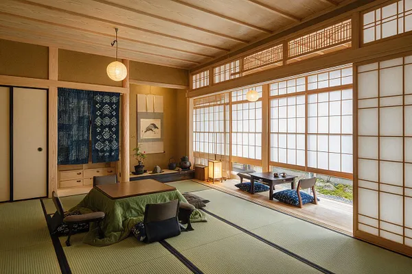 A Japanese Zen master bedroom with tatami mat flooring, a low table surrounded by floor cushions, and warm wood framing throughout. Shoji screens, paper lantern lighting, and a tokonoma-style alcove with a hanging textile and wall art create a calm, minimalist atmosphere.
