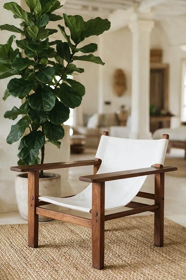 A tropical-style lounge chair with a dark wood frame and a white sling seat sits on a natural woven rug. A tall leafy potted plant stands beside it in a bright, neutral room with airy arches and soft beige decor.