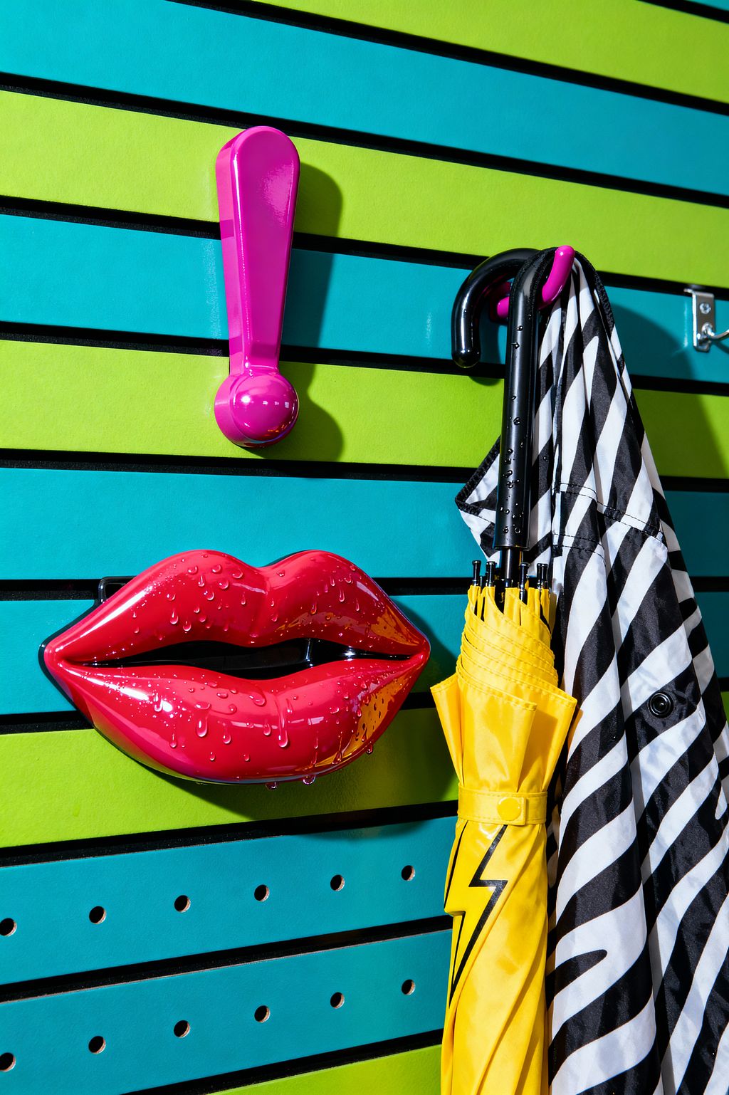 A mudroom wall with horizontal slatwall panels alternating neon lime green and teal with black grooves. A glossy hot-pink exclamation-point hook and an oversized pink lips hook, both shiny with water droplets, add a comic-book feel. To the right hang a black curved-handled umbrella, a folded yellow umbrella with a black lightning bolt, and a black-and-white diagonal striped raincoat on metal hooks.