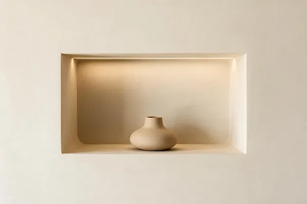 A minimalist wall niche painted in warm beige holds a small sculptural ceramic vase centered on the shelf. Soft recessed lighting washes the alcove, creating a calm, understated focal point against the smooth neutral wall.
