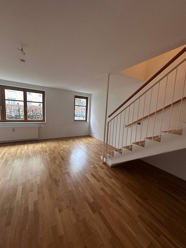 A spacious, empty room in an apartment with wooden flooring and a staircase.