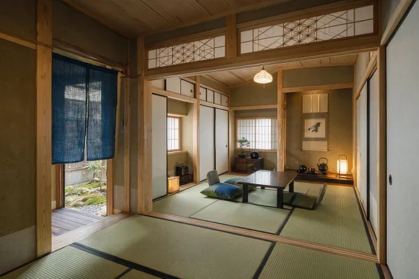 A Japanese Zen-style basement room with tatami mats, light wood framing, sliding shoji-style panels, and a low table surrounded by floor cushions. A blue noren curtain hangs at the entrance, while a tokonoma alcove with a hanging scroll and tea set glows under soft lantern lighting.
