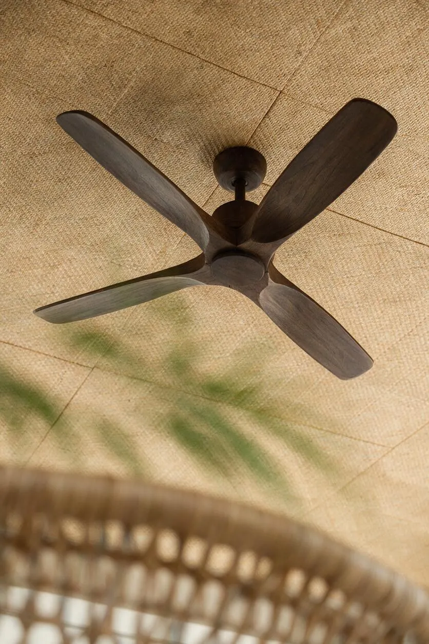A dark wood ceiling fan with four wide blades is mounted to a ceiling covered in a woven natural-fiber surface. Soft green leaf shadows drift across the warm tan background, and a blurred wicker pendant hangs in the lower foreground.