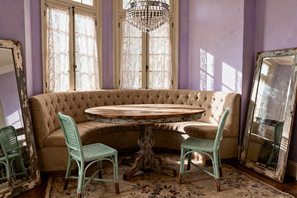 Dining nook with a curved tufted beige banquette surrounding a round distressed wood pedestal table, accented by two mint green rattan chairs. Lavender walls and tall French windows with lace curtains cast patterned light. A crystal-beaded chandelier hangs above, and two large leaning mirrors with chipped frames flank the seating. A faded floral rug lies over aged wood flooring.