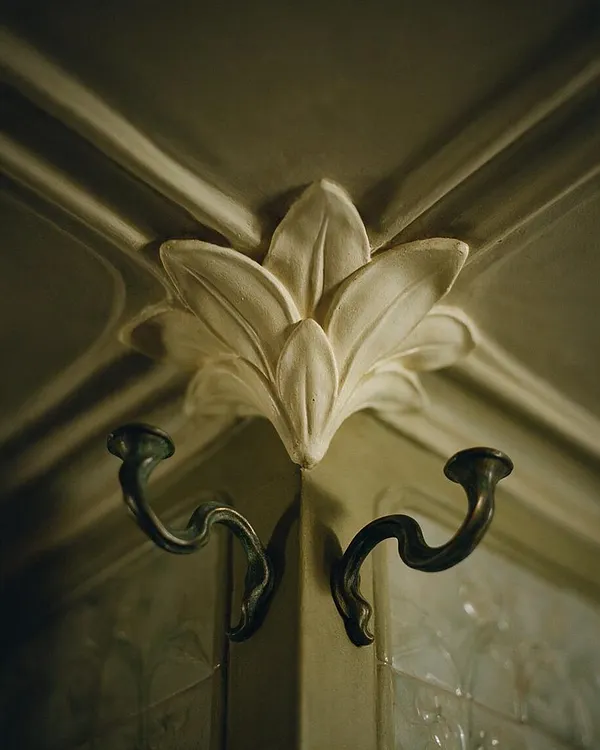 A close-up of an Art Nouveau interior detail showing a carved floral plaster ornament at the corner of a ceiling or wall junction. Beneath it are two dark, curved metal hooks with sinuous organic shapes, set against muted olive and beige surfaces with soft light and shadow.