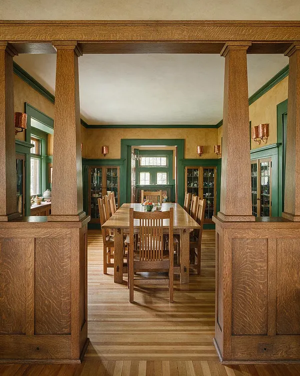 A Craftsman-style dining room framed by thick natural wood columns and trim, with a long wooden dining table and matching slat-back chairs centered on warm hardwood floors. Green window and door casings, glass-front built-ins, and copper wall sconces add rich color and period detail.