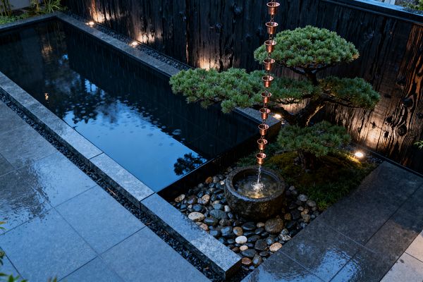 A minimalist Japanese-style patio features a rectangular reflecting pool edged with smooth gray stone, set beside a charred-wood privacy wall lit by ground uplights. A copper rain chain empties into a carved stone basin amid river pebbles and black gravel, while a sculpted pine and moss create a green focal point; wet pavers and gentle ripples from falling rain enhance the tranquil, evening atmosphere.
