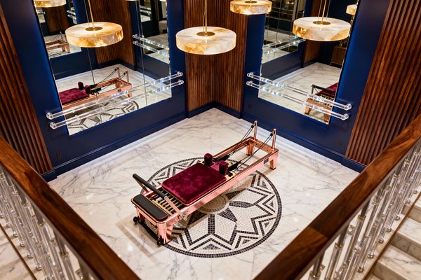 An elegant home gym features a rose-gold Pilates reformer with burgundy velvet pads centered on a circular black-and-white mosaic set into a white marble floor. Deep navy walls host large mirrors and clear acrylic ballet barres alongside vertical wood slat panels, while round stone-and-brass pendant lights glow above and an acrylic-and-brass stair balustrade lines the foreground.