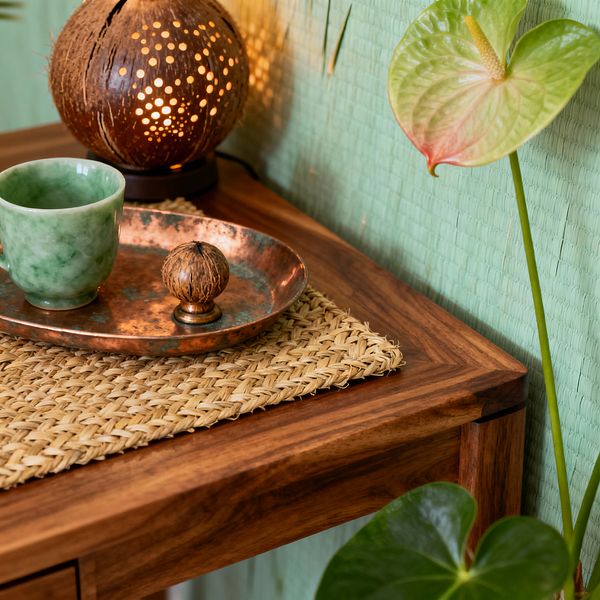 Close-up of a tropical home office corner with a solid teak desk topped by a braided seagrass mat. A hammered copper tray holds a marbled green ceramic cup and a small carved seed on a brass base. A perforated coconut-shell lamp casts warm dotted light on a mint-green grasscloth wall, while glossy leaves and an anthurium bloom frame the scene, emphasizing natural textures and an earthy palette.