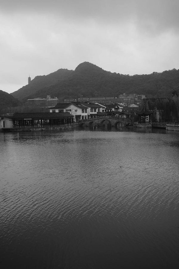 A tranquil lakeside scene at Xianghu Hu during sunset, featuring a calm water body, distant buildings, mountains, and a moored wooden boat with a person on it.