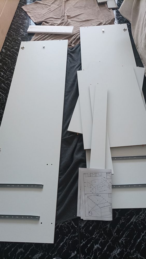 An open cardboard box containing flat-packed furniture parts, partially assembled, along with an instruction manual and some hardware items placed on the floor.