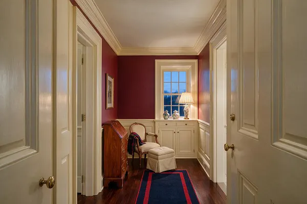 A traditional hallway or front entry space with deep burgundy walls, white crown molding, and paneled wainscoting. A small window with a lamp, upholstered chair, ottoman, and wooden secretary desk create a warm, elegant, lived-in feel.