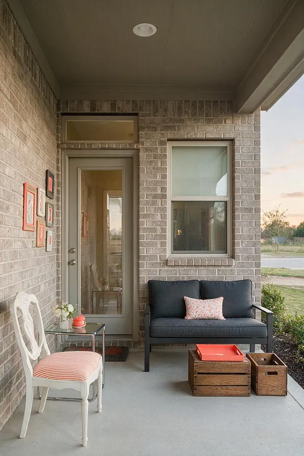 A covered front porch with gray brick walls and a taupe ceiling features a mix of seating styles, including a black upholstered loveseat, a white vintage chair with a striped cushion, and a small glass side table. Wooden crates and a coral tray add casual, eclectic charm against the soft neutral exterior.