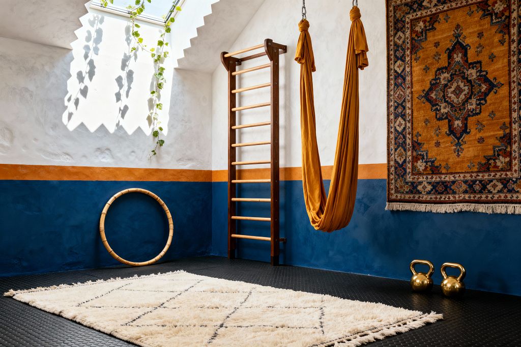 A sunlit home gym with a skylight and trailing vine features two-tone plaster walls: soft white above and deep indigo below, divided by an ochre stripe. A dark-wood stall bar and a mustard fabric aerial hammock anchor the space, with a Persian-style wall rug, a rattan hoop, and two brass kettlebells on black rubber flooring. A plush cream Moroccan shag rug softens the center, creating an airy, bohemian workout zone.