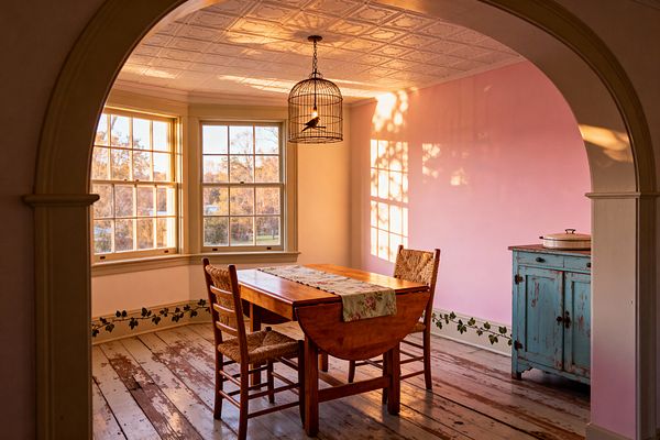 Viewed through an arched doorway, a small dining room features a warm wood drop-leaf table with two ladder-back chairs and a floral runner. Sunlight from multi-pane windows washes over dusty pink walls, cream trim, and a white pressed-tin ceiling. The space includes a distressed turquoise sideboard, a metal birdcage pendant light, a green ivy stenciled baseboard, and weathered white plank floors.