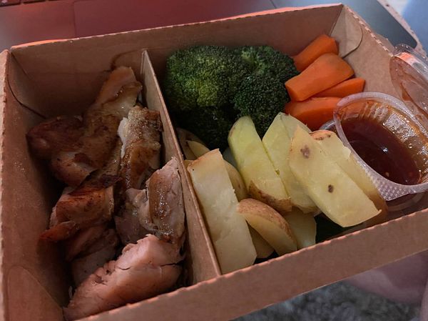 A meal box containing grilled chicken, vegetables, and dipping sauce.