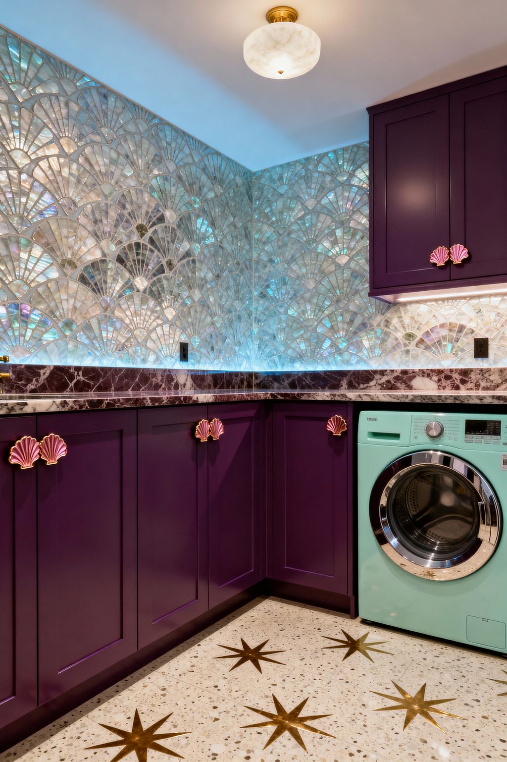 A vintage glam laundry room with L-shaped deep plum cabinets fitted with pink-and-brass shell pulls and a mint green front-load washer beneath a richly veined purple marble countertop. Iridescent fan-pattern glass tiles cover the walls and glow with under-cabinet LEDs, while a cream terrazzo floor with brass starbursts and a brass-and-alabaster ceiling light complete the jewel-box space.