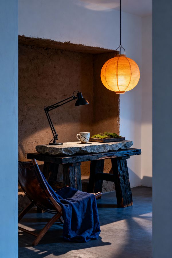 A compact home office sits within an earthen wall niche, featuring a rough stone slab desk on weathered dark timber trestles, lit by a warm orange paper lantern and a black adjustable task lamp. On the desk are a crackle-glazed ceramic mug and a shallow tray of moss, while a wooden-framed leather sling chair draped with an indigo linen throw rests on smooth concrete flooring with soft, moody shadows.