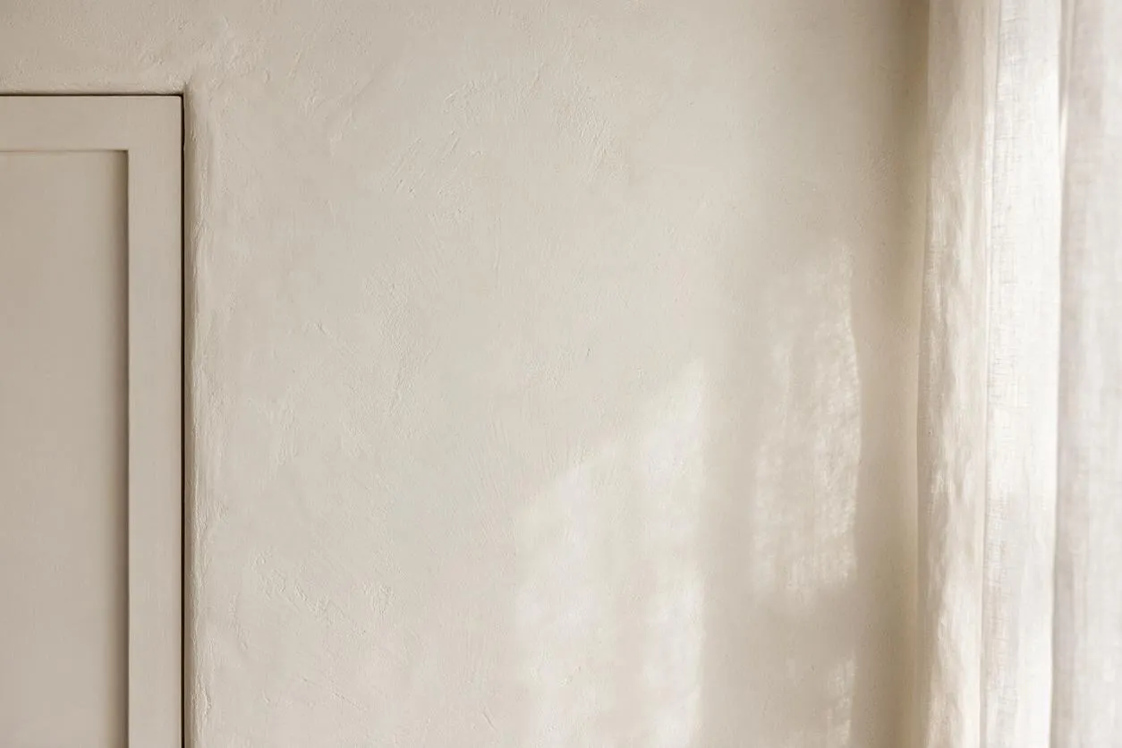 A close-up interior view of a softly textured beige plaster wall beside a simple framed panel on the left and sheer off-white curtains on the right. Natural daylight filters across the surface, creating a warm, calm Transitional look.