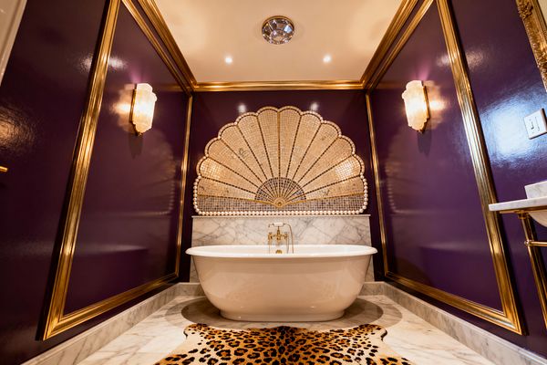 A glamorous bathroom with high-gloss purple walls framed in gold moulding, featuring a white freestanding tub below a scalloped fan mosaic made of gold and pearl tiles. Carrara marble covers the floor and low wainscot, alabaster sconces and a crystal flush-mount light cast warm illumination, and a leopard-print hide rug adds bold contrast.