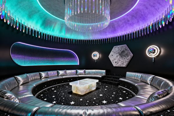 A circular media room features a sunken, curved sectional in metallic silver surrounding a glossy black floor scattered with white star inlays and a glowing alabaster coffee table. Matte black walls display a capsule-shaped LED panel, chrome ring sconces, and a hexagonal moon-textured art piece, while a domed ceiling with teal-to-violet cove lighting and an iridescent prism chandelier creates a futuristic, space-inspired atmosphere.