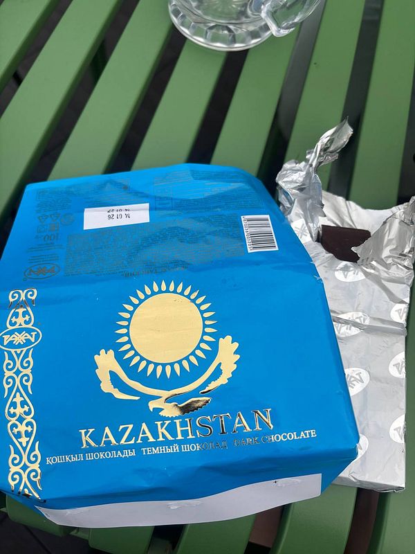 Blue packaging of Kazakhstan dark chocolate partially unwrapped on a green table next to a glass cup.