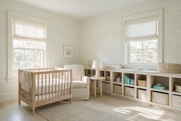 A bright coastal nursery with white shiplap walls, two tall windows with linen Roman shades, a light wood crib, a cream armchair, and a long built-in cubby unit filled with woven baskets and soft decor. Natural light washes over the pale wood floor and neutral rug.