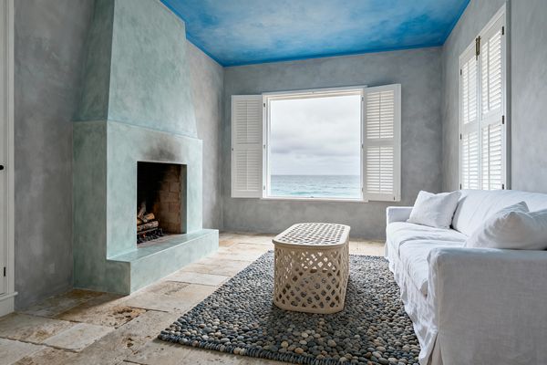 Coastal living room with a large window and white plantation shutters opening to an ocean view. A seafoam-green plaster fireplace sits on the left against limewashed gray walls, beneath a sky-blue ombré ceiling. On the right, a white linen slipcovered sofa faces a pebble-textured gray rug topped with an oval woven rattan coffee table. The floor is warm travertine tile, and soft natural light creates a calm, breezy atmosphere.