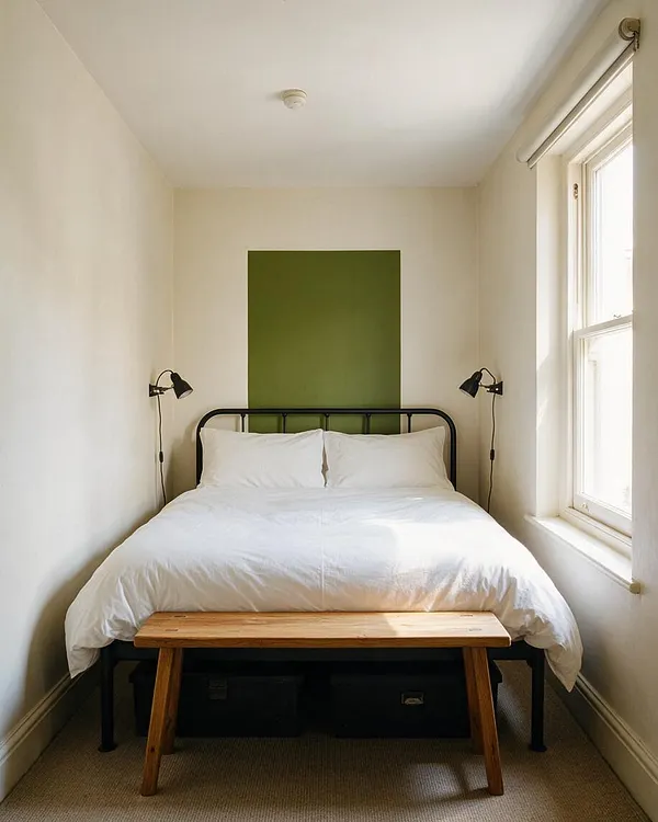 A narrow modern guest bedroom with a black metal bed frame dressed in white bedding, centered against a plain white wall with a vertical olive-green accent panel behind the headboard. Two black wall-mounted reading lamps flank the bed, a simple wooden bench sits at the foot, and a bright window on the right brings in natural light.