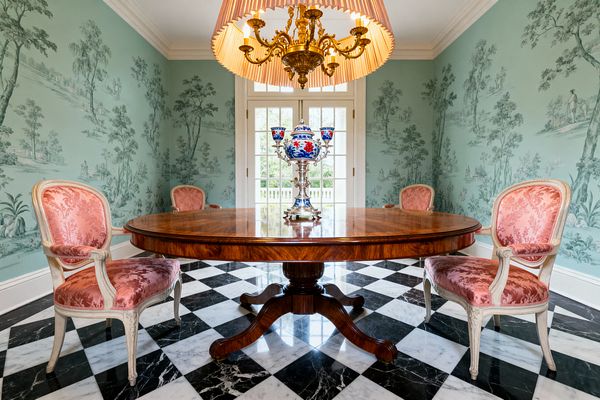 A traditional dining room with a round mahogany pedestal table centered on a black-and-white marble checkerboard floor. Four Louis-style armchairs with cream-painted frames and pink damask upholstery surround the table, against mint-green walls in scenic toile. A gilt chandelier with a pleated drum shade hangs above, and divided-light French doors brighten the space. A blue-and-red porcelain centerpiece sits on the tabletop.