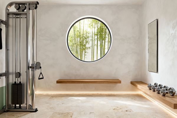 A minimalist home gym with warm white plaster walls features a large circular window centered on a view of bamboo. Creamy stone tiles cover the floor. Floating oak benches line two walls; one supports a row of chrome dumbbells. A slim rectangular mirror hangs on the right, and a stainless-steel cable machine stands on the left. Concealed LED strip lighting runs along the base and under the benches, casting a soft glow that emphasizes clean lines and open space.