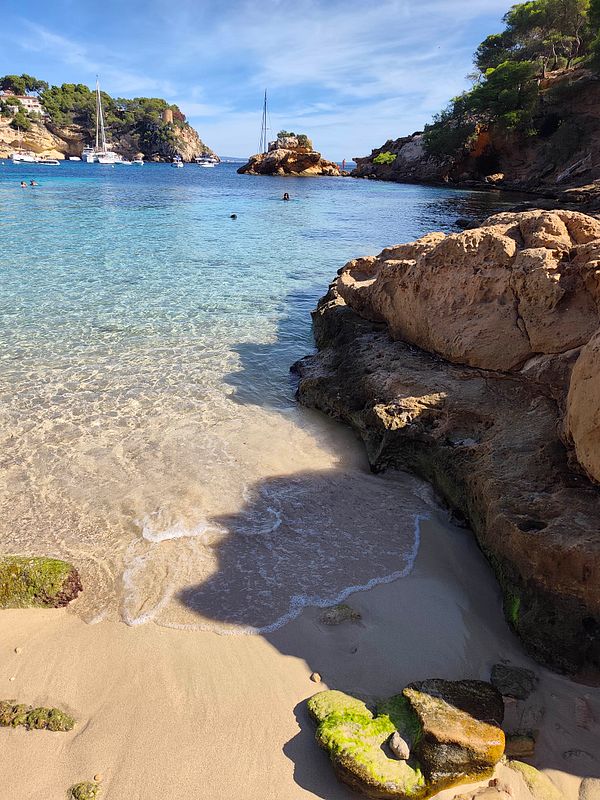 A serene coastal scene with clear blue water, rocky cliffs, a sandy beach, and several people swimming and sailboats anchored in the distance.