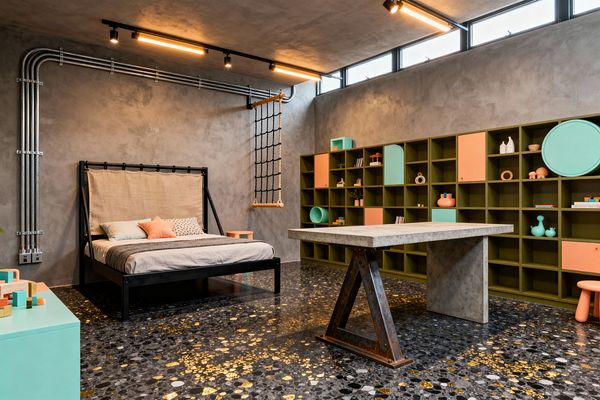 Industrial-themed kids’ room with gray concrete walls, clerestory windows, and exposed metal conduit. A black metal bed with a canvas headboard sits beside a hanging rope ladder, opposite a long olive-green shelving wall with mint and coral cabinet fronts. In the center, a cast-concrete table with a steel trestle stands on black terrazzo flooring dotted with gold flecks. Warm linear track lights illuminate toys and playful accents.