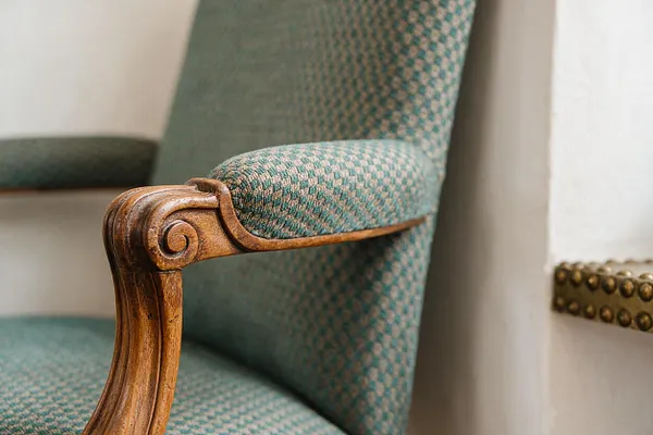 A close-up view of a vintage upholstered armchair with a carved wooden armrest and teal patterned fabric. The chair sits beside a light wall with a small section of brass nailhead trim visible in the background, highlighting the mix of classic detailing and textured fabric.