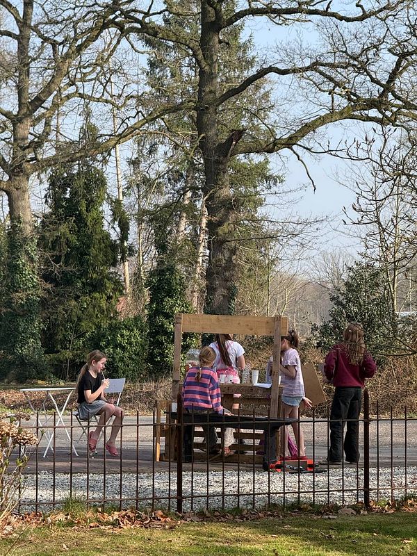 Two children running outside on a paved path holding lemonade cups with straws, with a grassy green field and a wooden fence in the background.