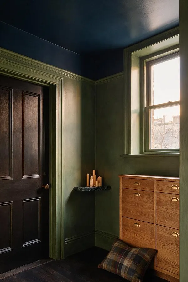 A dark academia entryway with deep green walls, a dark wood paneled door, and a warm oak drawer cabinet beneath a bright window. A small black wall shelf holds candles, and a plaid floor cushion adds a scholarly, lived-in feel.