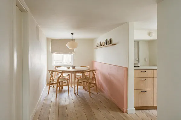 A narrow basement dining area with pale wood plank floors, white walls, and a round light wood table surrounded by matching Scandinavian chairs. A blush pink half-wall runs along one side, with a simple pendant light overhead, a linen window shade, and a small kitchen alcove visible to the right.