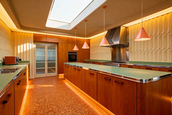 A long Mid-Century Modern kitchen with a central island shows mint-green laminate countertops with chrome edging over warm wood cabinets, sculpted cream tile walls, and a stainless steel range with an oversized hood. Coral cone pendants, cork flooring, warm cove and toe-kick lighting, a double sink with small appliances on the left, and ribbed-glass sliding doors complete the space.