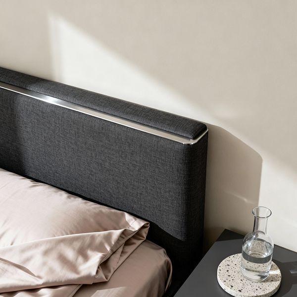 A close-up of a minimalist bedroom corner showing a dark charcoal upholstered headboard with a slim polished metal trim along the top, set against a warm off-white wall. Soft blush-taupe sheets drape neatly over the mattress. Beside the bed, a matte graphite nightstand holds a clear glass carafe resting on a round terrazzo coaster. Natural daylight streams in from the left, casting crisp geometric shadows that emphasize clean lines and uncluttered surfaces.