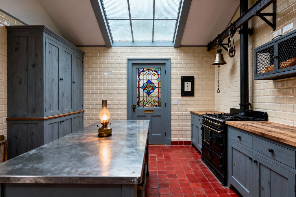Victorian-style kitchen with a large skylight, cream beveled subway-tile walls, and a red quarry-tile floor. Slate-blue painted cabinets with black knobs and butcher-block counters line the right wall around a black cast-iron range and exposed flue; a pulley rail with bell and a wire-mesh wall cupboard sit above. A tall pantry and a zinc-topped island with an oil lamp stand to the left, and a blue-gray door with colorful stained glass centers the far wall.