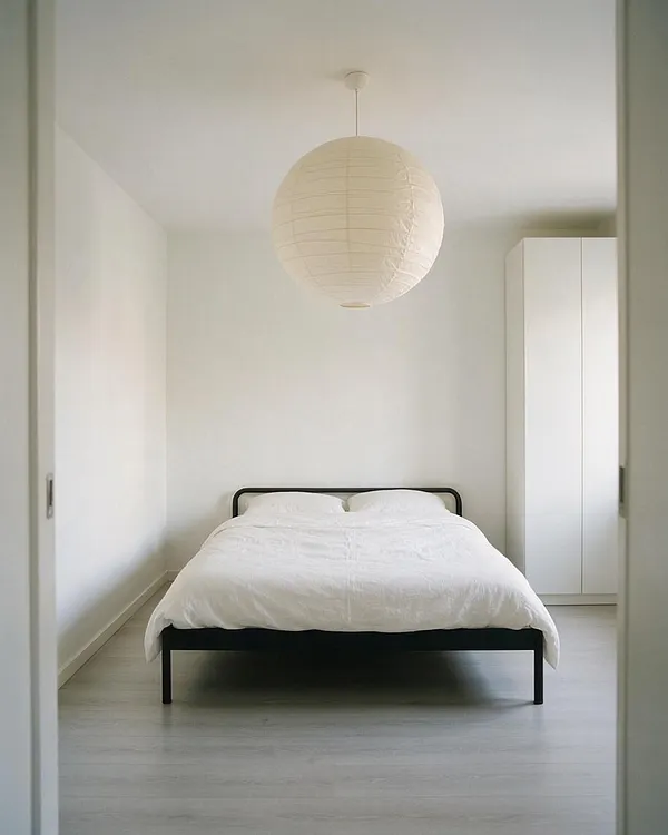 A minimalist master bedroom with white walls and light flooring features a low black metal bed frame dressed in crisp white bedding. A large round paper lantern hangs from the ceiling, and a tall white wardrobe stands against the right wall.