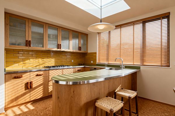 A mid-century kitchen features a rounded peninsula with olive-green laminate and chrome edging, oak cabinets with sculpted wood pulls, and upper cupboards fitted with ribbed glass. Glossy mustard subway tiles line the backsplash, a stainless-steel sink and chrome faucet sit beneath wide wooden blinds and a skylight, and a saucer pendant, gas cooktop, cork flooring, and two woven-seat stools complete the warm, sunlit space.