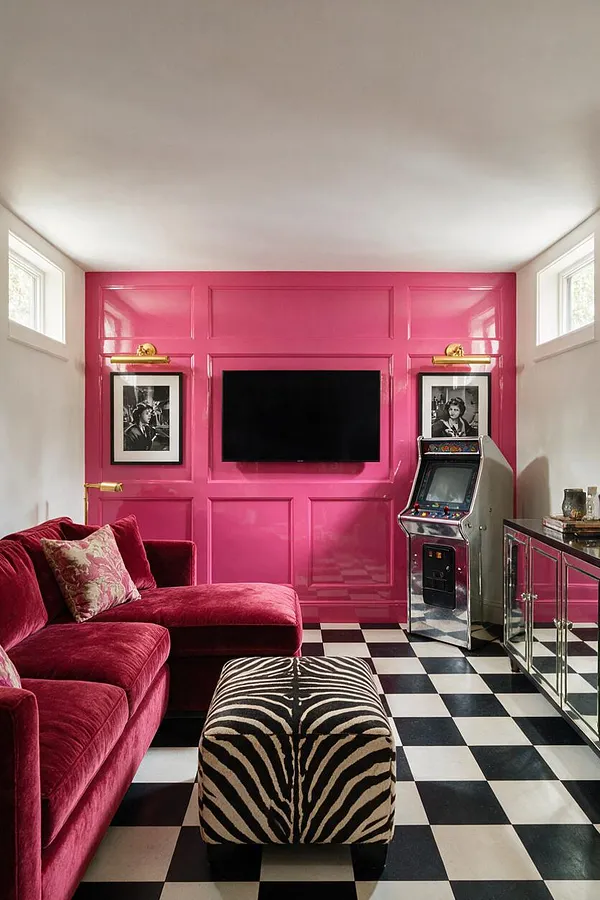 A dramatic game room features a glossy hot pink paneled accent wall with a wall-mounted TV, black-and-white checkered flooring, and mirrored cabinetry. A deep burgundy velvet sectional and zebra-print ottoman add bold texture, while small framed black-and-white portraits and a vintage arcade cabinet reinforce the glamorous, playful Hollywood Regency style.