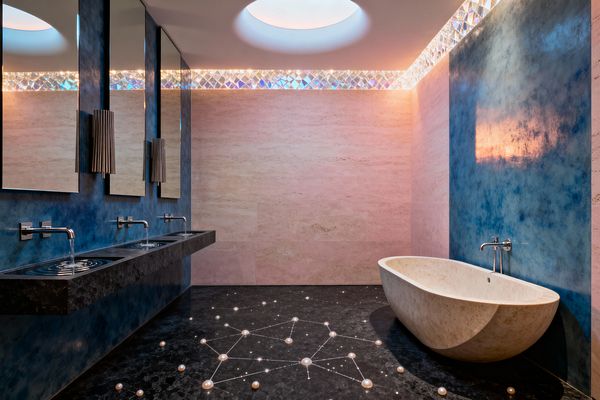 A spacious bathroom features blush travertine walls and a deep blue plaster accent. On the left, a floating black stone vanity with three round basins sits beneath rectangular mirrors and ribbed sconces, with chrome wall-mounted faucets. On the right, an oval limestone freestanding tub rests on black marble flooring inlaid with constellation-like lights and pearl-like spheres. A circular ceiling opening and a band of faceted glass cast soft, iridescent illumination.