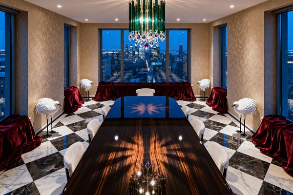 A glamorous dining room features a glossy dark-wood rectangular table centered beneath an emerald-glass and crystal chandelier, its facets reflected on the tabletop. The room has a black-and-white checkerboard marble floor, cream upholstered chairs, champagne textured walls, and tall windows overlooking a nighttime city skyline; burgundy velvet–draped consoles run beneath the windows, flanked by white bird sculptures on black stands, with recessed ceiling lights adding soft glow.
