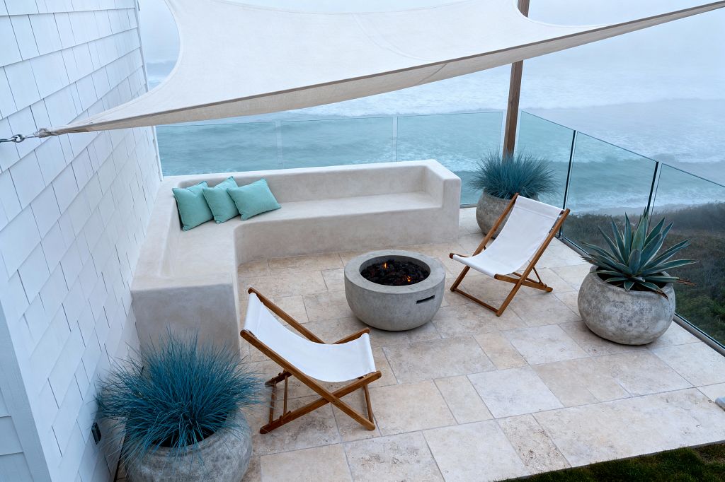 An oceanfront patio features an L-shaped built-in bench in smooth light-beige stucco beside a glass balustrade overlooking waves. A round concrete fire pit anchors the space, flanked by two white canvas sling chairs with wooden frames on beige travertine pavers. A triangular sand-colored shade sail stretches overhead. Large weathered concrete planters hold blue fescue grass and a sculptural agave, while three seafoam pillows rest on the bench.
