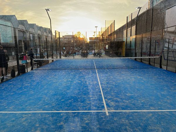 A vibrant outdoor padel court at sunset, surrounded by people and modern buildings.
