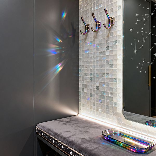 Close-up of a Cosmic Chic mudroom nook with matte charcoal cabinetry, a gray velvet bench seat edged with star studs and nailhead trim, and a wall of pearlescent glass mosaic illuminated by a concealed LED strip. Three iridescent metal hooks sit beside a beveled mirror with constellation graphics, while a rainbow-glass tray rests on the bench and light casts prismatic reflections.