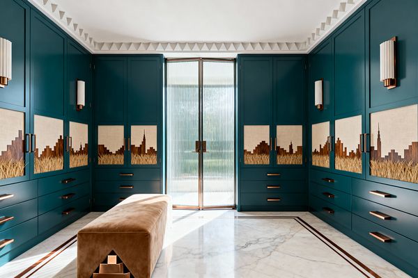 A symmetrical mudroom is lined with floor-to-ceiling teal cabinets whose linen-backed walnut marquetry panels depict a skyline rising above wheat, accented by brushed copper pulls and fluted white sconces. A camel velvet bench with a stepped copper detail rests on a white marble floor with a bronze inlay border, while reeded-glass double doors admit daylight beneath a stepped geometric ceiling cornice.