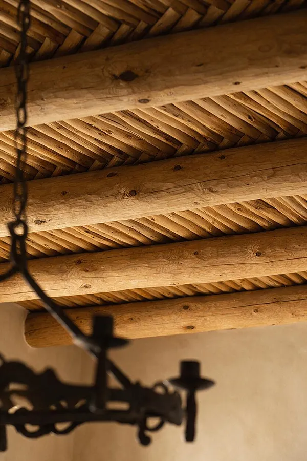 A close-up view of a Southwestern-style ceiling showing thick exposed log beams and a woven reed or branch infill pattern between them. A dark wrought-iron light fixture hangs out of focus in the foreground against a warm beige wall.